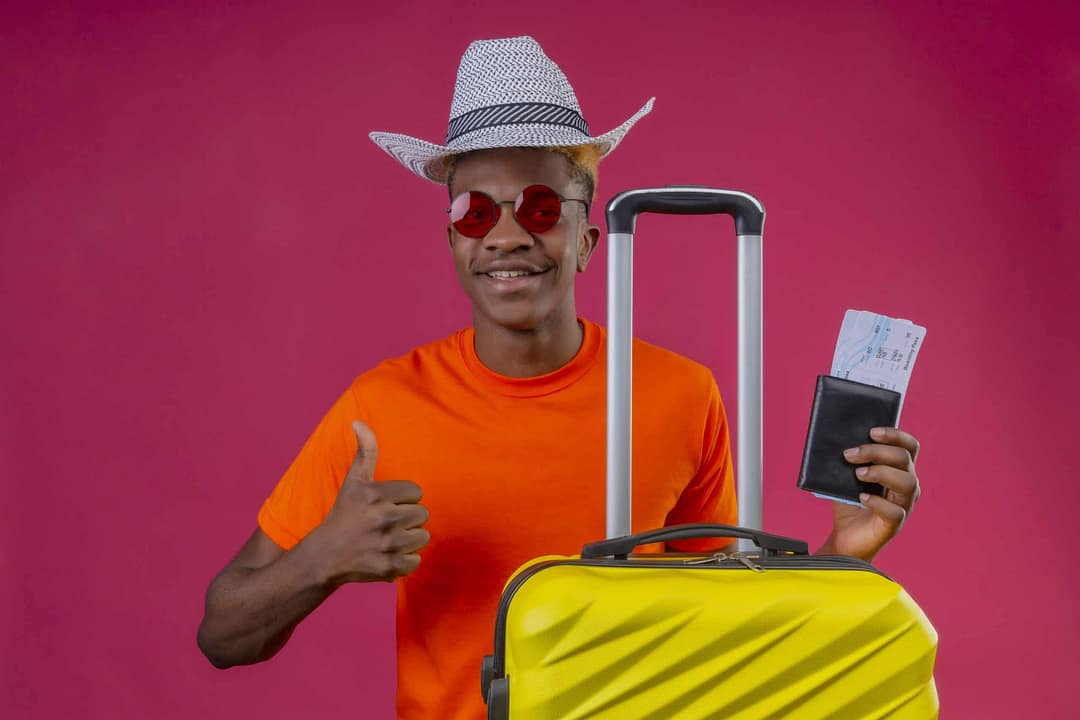 young african american traveler boy wearing orange t-shirt and summer hat holding travel suitcase and air tickets looking at camera smiling positive and happy showing thumbs up standing over pink background